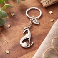Close-up of a silver greyhound/whippet keyring on a warm wood surface. The keychain is styled with rustic elements including a twine heart, scattered gold stars, green sprigs, and a textured linen cloth, creating a cozy and artisanal feel