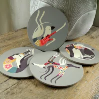 Greyhound and lurcher coaster set displayed on a wooden table, showing four round coasters with stylised sighthound illustrations on a soft grey-green background. Each coaster features a different pose, including stretching, running, resting, and curling greyhounds and lurchers, with a textured vase in the background creating a cosy home setting.