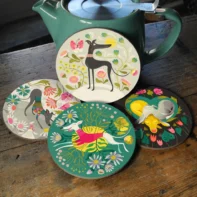 Happy Hounds Coaster Set displayed on a rustic wooden table, featuring four round illustrated coasters with stylized greyhounds and floral designs. The coasters show different scenes: a stretching greyhound, a leaping greyhound in a pink sweater, two curled greyhounds resting, and a standing greyhound surrounded by flowers. A green teapot sits behind the coasters, adding a cosy, home setting.